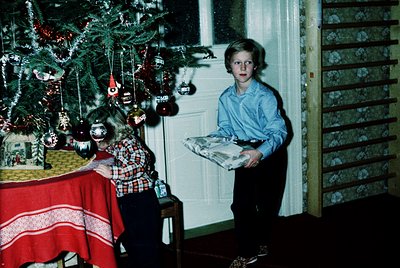 Vintage 1970s holiday scene featuring two children in a modestly decorated living room. A boy in a blue shirt holds a wrapped...