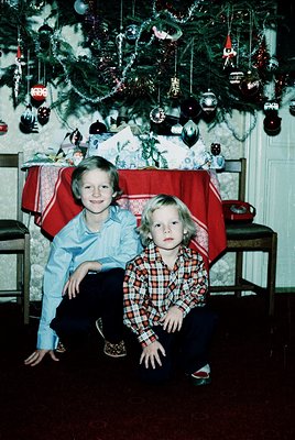 Two children pose under a festively decorated Christmas tree in a 1970s-style interior. The boy kneels in a plaid shirt and d...