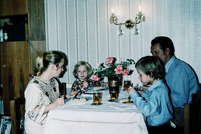 Family dinner scene in a mid-century interior, featuring four individuals seated around a table adorned with floral centerpie...