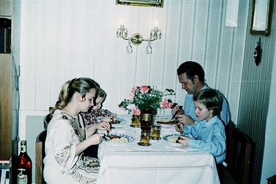 Family meal scene in a mid-century interior, featuring three children and an adult seated around a round table. The table is ...