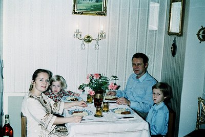 Family portrait from the 1970s–1980s, seated at a vintage dining table with floral centerpiece. Mother wears embroidered blou...