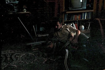 Darkly lit room featuring a man in military attire kneeling beside a laptop on a patterned carpet. Surrounding him: vintage C...