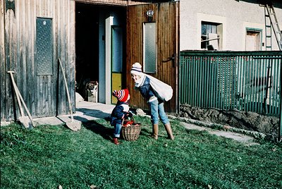 A woman in 1970s-era denim and headscarf assists a child in a striped sweater and red shoes with a woven basket, likely gathe...