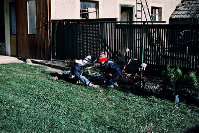 Two children play with a small orange lawnmower in a residential yard, 1970s-80s. Whitewashed house with wooden door and blac...