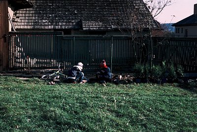 Vintage-style photo of two individuals resting on grass beside a fallen bicycle in a residential backyard. Wooden fence with ...