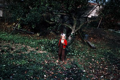 Vintage black-and-white photo of a child standing amidst fallen autumn leaves under a large tree. The child wears a striped s...