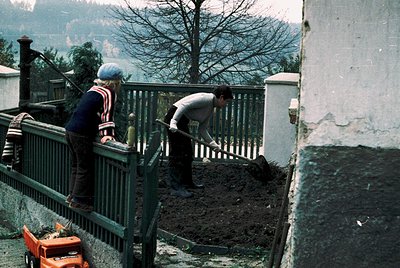 Two men in 1970s-era workwear shovel dirt beside a weathered concrete wall and metal railing, likely for construction or land...