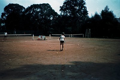Vintage tennis court with clay surface, surrounded by trees and a fence. A lone player in a striped shirt and shorts prepares...