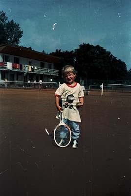 Vintage tennis court scene featuring a young boy in a white T-shirt (with "G" logo) and rolled-up jeans, holding a red-and-wh...