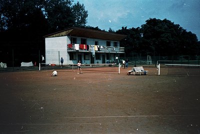 Mid-century tennis court with red clay surface, bordered by white lines. Two players in motion near net; spectators seated on...