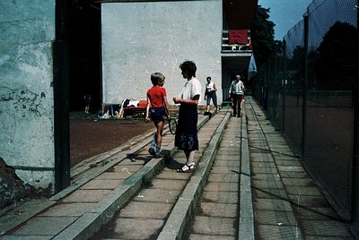 Vintage urban courtyard with worn concrete steps and brick walkway. A woman in a white blouse and dark skirt leads a child in...