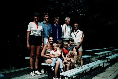 Family group shot in outdoor stadium seating, likely from the 1970s–1980s. Eight individuals pose on wooden bleachers under s...