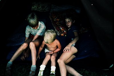Three children sit on a blue inflatable pool in a dimly lit backyard, likely mid-1970s. The boy on the left wears a striped s...