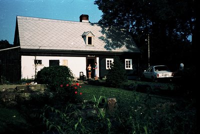 Single-story rural house with gabled roof and whitewashed walls, framed by overgrown garden beds. A person stands in the door...