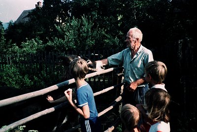 A man in a light-colored shirt and dark pants interacts with children at a zoo enclosure, pointing toward a brown bear behind...