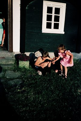 Vintage black-and-white photo capturing a candid moment: a woman in a sleeveless dress kneels beside a young girl in a pink d...