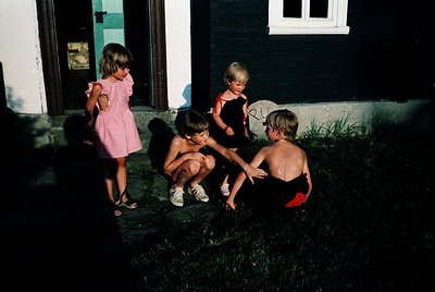 Four children play in a sunlit backyard, 1960s-1970s. Girl in pink dress kneels beside boys in shorts, one shirtless. Whitewa...