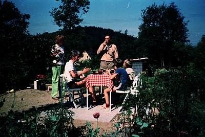 Vintage outdoor gathering in a lush garden setting, featuring four adults seated at a red-and-white checkered table. Central ...