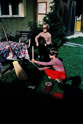 Two children in 1960s-style striped swimwear play with a bamboo pole and bucket in a residential backyard. The boy kneels, ho...