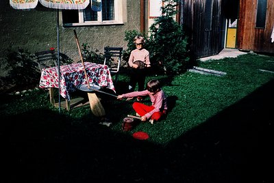 Two children play on a grassy backyard lawn, 1960s-70s. Child on left kneels in red pants, holding a toy rake; child on right...