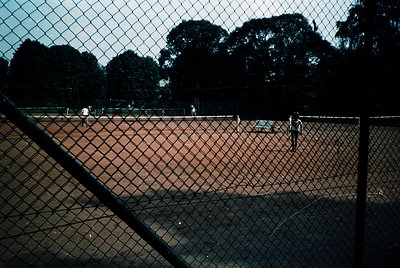 Vintage clay-court tennis match framed by chain-link fence. Players in motion, lush green trees in background. Likely mid-20t...