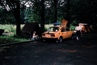 Vintage orange hatchback car parked beside open camping tents in a wooded area, likely a 1970s-80s European campsite. Two adu...