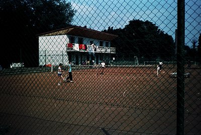 Vintage tennis court with chain-link fence, featuring a two-story building in background. Players in retro attire (1970s-80s)...