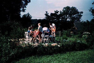 Vintage family gathering on a rustic wooden deck surrounded by lush greenery, likely mid-20th century. Five individuals seate...