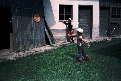 Vintage photo of two children playing in a courtyard with weathered, rustic buildings. Child in red shoes chases another hold...