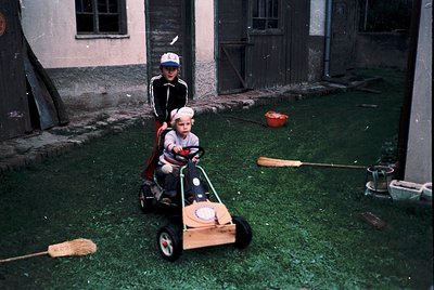 Two children play in a courtyard with mid-century toy cars, one with a steering wheel. The older child wears a striped shirt ...