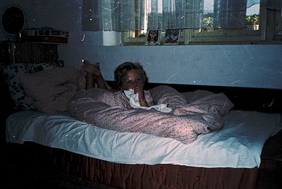 Vintage interior shot of a child in a nightgown resting on a twin bed under a window with lace curtains. Wooden headboard and...