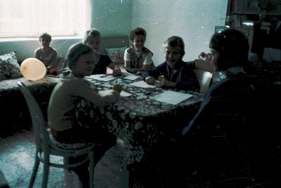 Vintage indoor scene of six adults playing checkers around a patterned tablecloth, likely mid-20th century. Indoor lighting a...