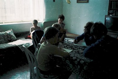 Vintage indoor scene featuring a group of four adults and a child gathered around a patterned tablecloth, likely for a meal o...