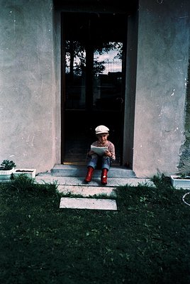 Child in red rubber boots and white cap sits on concrete steps reading a book, framed by a weathered gray door. Mid-20th cent...