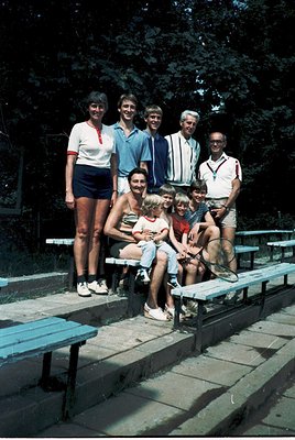 Family group shot in outdoor stadium seating, likely 1970s. Six adults and two children pose on concrete bleachers under shad...