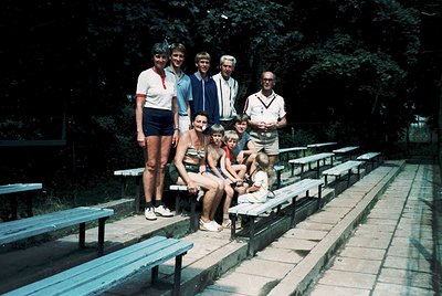 Family group shot in 1970s outdoor stadium seating, likely for sports or events. Eight individuals pose on tiered concrete bl...