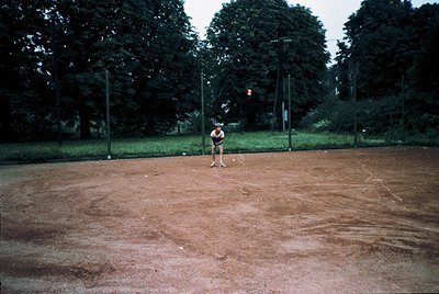 Baseball player in mid-action on a dirt infield, shadowed by dense trees. Classic wooden fence and numbered posts (, ). Likel...