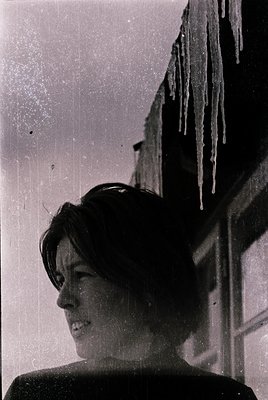Black-and-white portrait of a woman with short, dark hair, captured mid-smile, standing under icicles dripping from a buildin...