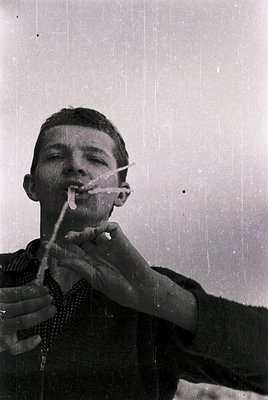 Vintage black-and-white portrait of a young man holding a cigarette between his lips while adjusting a striped necktie. Mid-2...