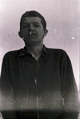 Vintage black-and-white portrait of a young man in a checkered shirt, likely mid-20th century. His direct gaze and slight smi...