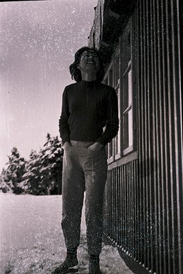 Black-and-white street scene: Young person in 1970s-style track pants and turtleneck stands under heavy snowfall, leaning aga...
