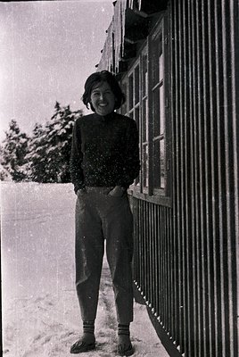 Young woman in 1960s winter attire—turtleneck sweater, wide-legged trousers, and knee-high socks—poses outdoors near a wrough...