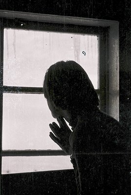 Silhouette of a person leaning against a window frame, hands clasped near face, framed by a single-pane glass window with vis...