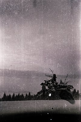 Vintage black-and-white ski lift ascending a forested slope, likely mid-20th century. Rustic wooden chairs carry skiers throu...
