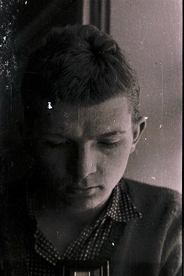 Vintage black-and-white portrait of a young man with a serious expression, captured in mid-20th century style. His hair is ne...