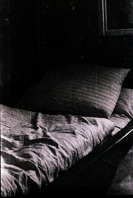 Unmade bed with rumpled sheets and a single pillow, minimalist interior with dark walls. Low-key lighting highlights texture....