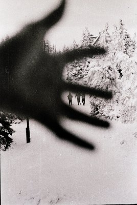 Vintage black-and-white photo of a shadow cast by a person’s arm and hand on snow, with three figures hiking in a forested al...