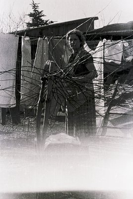 Mid-20th century black-and-white photo of a woman operating a manual clothes wringer, likely in a residential backyard. Barbe...