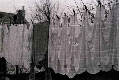 Mid-20th century black-and-white photo of laundry hung on a backyard clothesline. Checkered tablecloths and sheer curtains do...