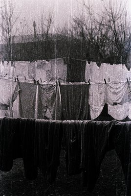 Mid-20th century laundry line with heavy cotton sheets, blankets, and garments hung to dry. Rustic wooden fence and bare tree...